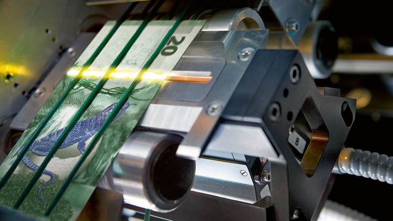 Close-up image of a banknote processing machine with ultrasonic sensors and a sample note