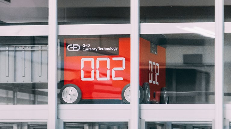 Behind the window pane of a high-rise building stands a red machine on wheels with the G+D Currency Technology logo 