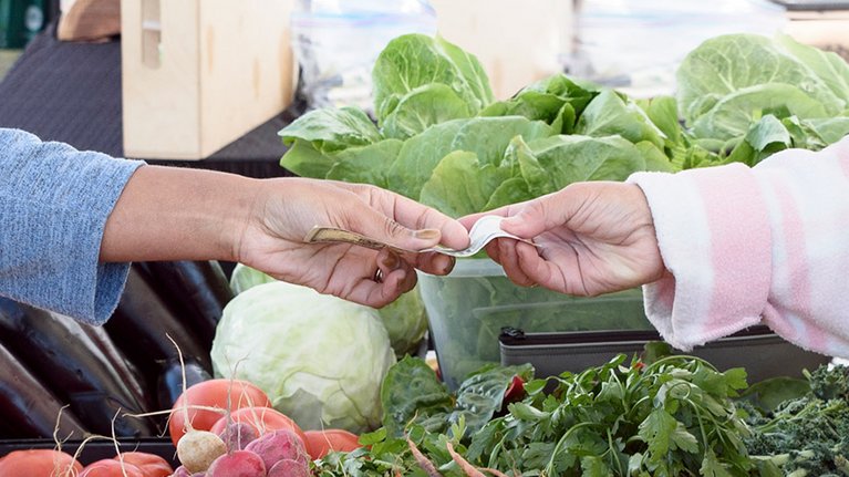 An einem Gemüsestand wird ein Geldschein zum Bezahlen überreicht