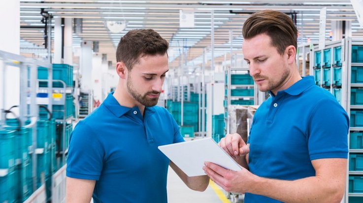 Two employees in blue polo shirts discuss a clipboard, NotaTrays are in the background